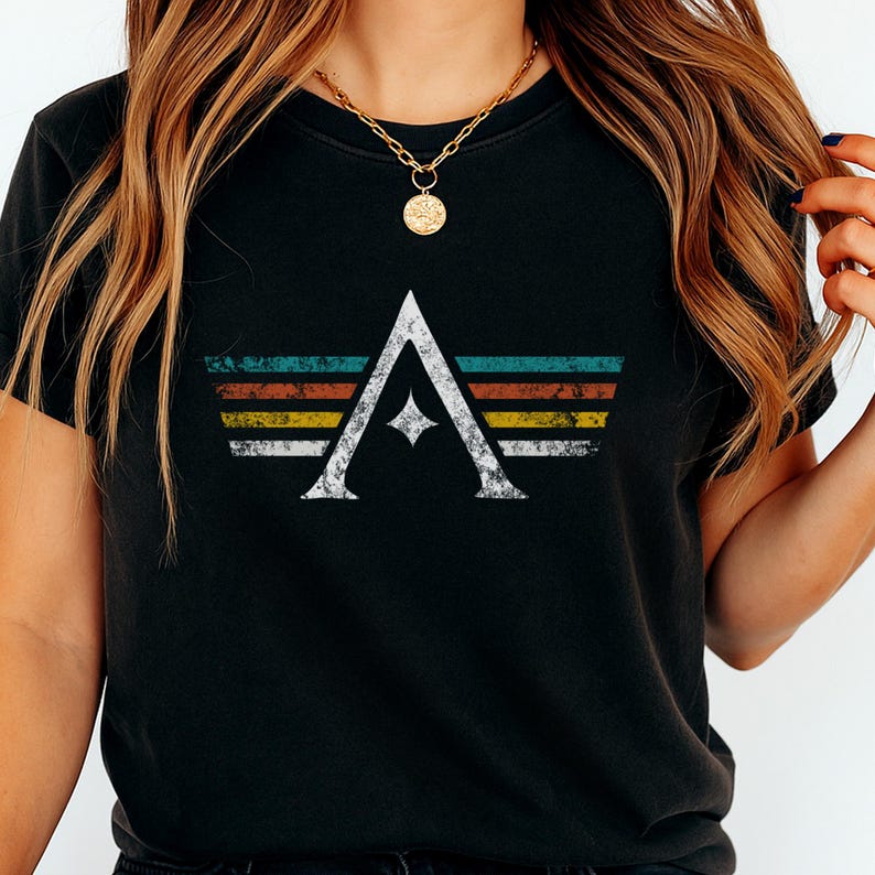 A minimalist t-shirt featuring the Arcs board game logo in a retro style. The design includes white geometric lines with a central star and vintage-colored stripes (teal, orange, red, yellow) on a dark background. Perfect for board gamers, sci-fi fans, and anyone who loves stylish and unique game night apparel.
