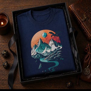Navy DnD mountain adventure shirt folded in a dark moody gift box — vintage fantasy landscape design as a tabletop RPG gift for dungeon masters and outdoor adventure fans