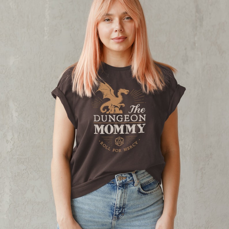 Dark red Dungeon Mommy T-shirt with a bold vintage-style dragon graphic above the phrase “Dungeon Mommy.” A large D20 dice and the words “Roll for Mercy” appear below, making this a funny and powerful gift for any Dungeons and Dragons fan. Ideal for gamer moms, dungeon masters, or fantasy lovers who want to show off their RPG pride in style. Perfect for birthdays, Mother’s Day, or any tabletop night.