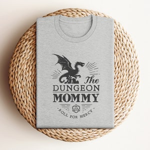 Dark red Dungeon Mommy T-shirt with a bold vintage-style dragon graphic above the phrase “Dungeon Mommy.” A large D20 dice and the words “Roll for Mercy” appear below, making this a funny and powerful gift for any Dungeons and Dragons fan. Ideal for gamer moms, dungeon masters, or fantasy lovers who want to show off their RPG pride in style. Perfect for birthdays, Mother’s Day, or any tabletop night.