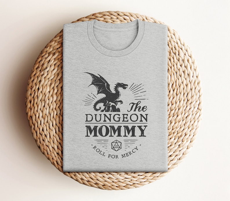 Dark red Dungeon Mommy T-shirt with a bold vintage-style dragon graphic above the phrase “Dungeon Mommy.” A large D20 dice and the words “Roll for Mercy” appear below, making this a funny and powerful gift for any Dungeons and Dragons fan. Ideal for gamer moms, dungeon masters, or fantasy lovers who want to show off their RPG pride in style. Perfect for birthdays, Mother’s Day, or any tabletop night.