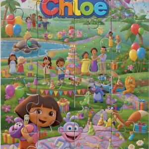 May include: A colorful jigsaw puzzle featuring a birthday party scene with Dora the Explorer and Boots. The puzzle, titled "Chloe," includes balloons, a cake, and various cartoon characters in a vibrant outdoor setting. The puzzle is made of multiple pieces.