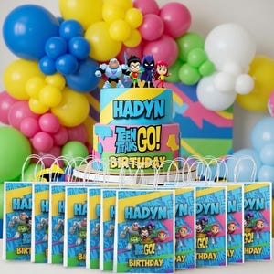 May include: A vibrant birthday party setup with a cake decorated with Teen Titans Go! characters and the name "Hadyin." Balloons in blue, yellow, pink, and green are arranged around the cake. Gift bags with the same theme are in the foreground.