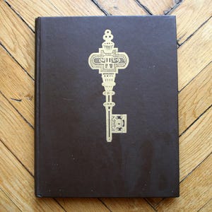May include: A dark brown hardcover book with a gold key design on the front cover.