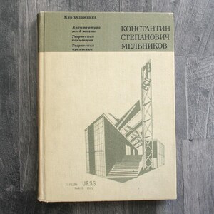 Puede incluir: Un libro antiguo con una cubierta verde claro y beige. La cubierta presenta texto en ruso y una ilustración arquitectónica geométrica en verde. El título del libro es "Konstantin Stepanovich Melnikov."