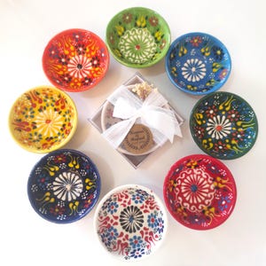 Op de afbeelding: Een verzameling van acht kleine keramische kommen met ingewikkelde bloemmotieven in rood, groen, blauw, geel en wit, gerangschikt rond een centrale vierkante geschenkdoos met een wit lint en een label met de tekst Karina & Rafael 11.04.2024.