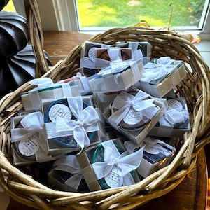 May include: A wicker basket filled with small, square gift boxes tied with white ribbon. Each box contains a small, colorful ceramic dish with a white sticker that says "Celebrating Gabriel 12.18.2024 With Love".