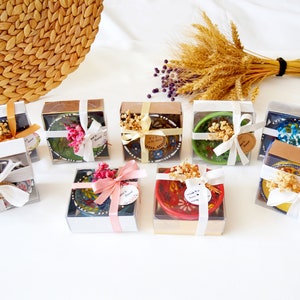 Op de afbeelding: Een assortiment van kleine, kleurrijke keramische kommen in geschenkdozen, gebonden met wit lint en versierd met gedroogde bloemen. De dozen zijn gerangschikt op een wit oppervlak.