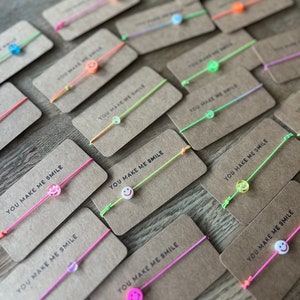 Könnte beinhalten: Eine Sammlung bunter Freundschaftsarmbänder mit Smiley-Anhängern. Jedes Armband ist an einer braunen Karte mit dem Text "You Make Me Smile" gebunden.