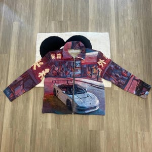 Peut inclure: Une veste zippée rouge et noire avec une voiture blanche et du texte japonais imprimé sur le devant. La voiture roule dans une rue avec des bâtiments de chaque côté.