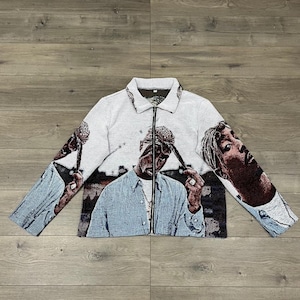 Puede incluir: Chaqueta blanca con cremallera con un diseño de tapiz con imágenes de una persona. La chaqueta tiene cuello y mangas largas. El diseño incorpora tonos de azul, marrón y negro.