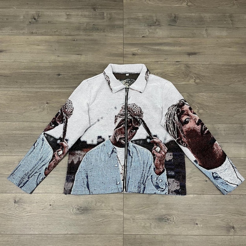 Op de afbeelding: Een witte jas met ritssluiting met een tapijtontwerp met afbeeldingen van een persoon. De jas heeft een kraag en lange mouwen. Het ontwerp bevat tinten blauw, bruin en zwart.