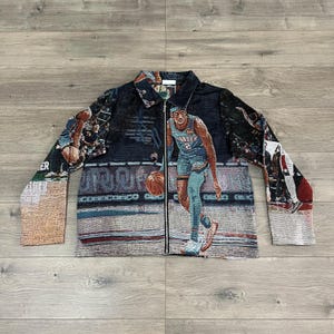 May include: A full-zip jacket with a basketball theme. The jacket features a woven design with a basketball player in action, with the word "THUNDER" visible. The jacket has a black background with blue, red, and tan accents.