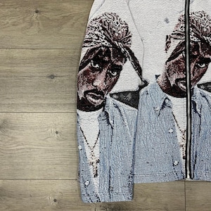 Op de afbeelding: Een wandtapijtjas met een portret van een persoon die een bandana en een spijkeroverhemd draagt. De jas heeft een zwarte ritssluiting en een lichtblauw en wit kleurenschema. Het portret is in tinten bruin, zwart en wit.