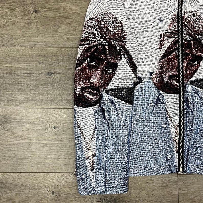 Op de afbeelding: Een wandtapijtjas met een portret van een persoon die een bandana en een spijkeroverhemd draagt. De jas heeft een zwarte ritssluiting en een lichtblauw en wit kleurenschema. Het portret is in tinten bruin, zwart en wit.