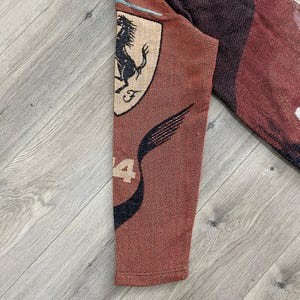 May include: A section of a brown textile featuring the Ferrari logo, a black prancing horse on a yellow shield, and the number 4. The textile has a woven texture and a dark red and black design.