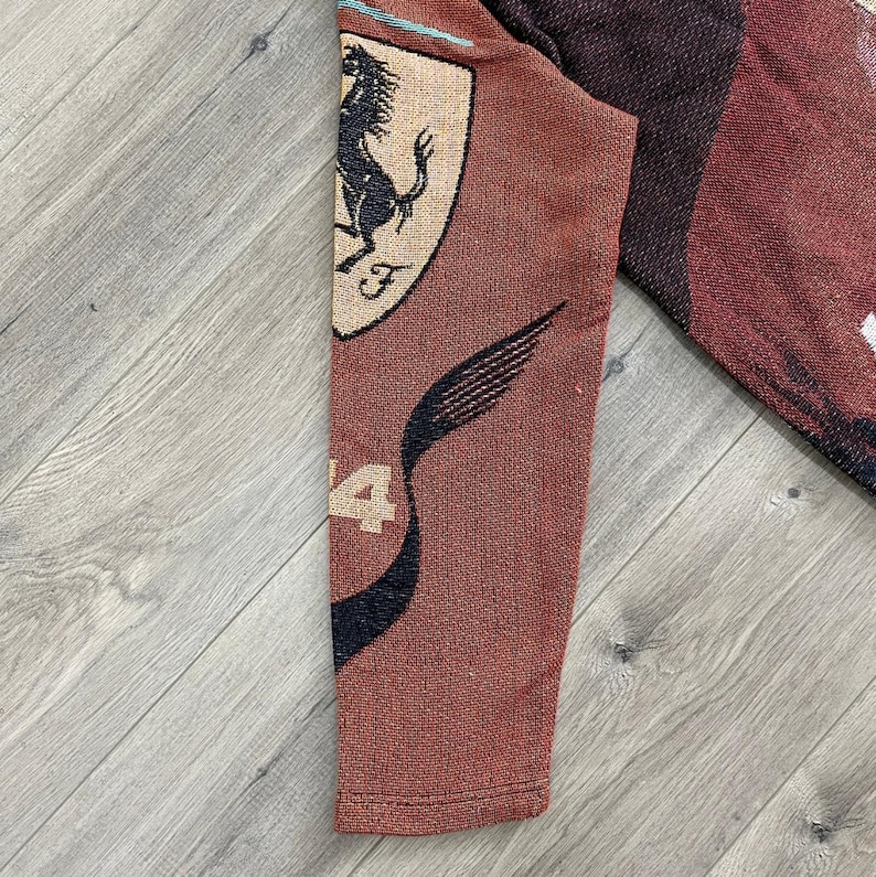 May include: A section of a brown textile featuring the Ferrari logo, a black prancing horse on a yellow shield, and the number 4. The textile has a woven texture and a dark red and black design.