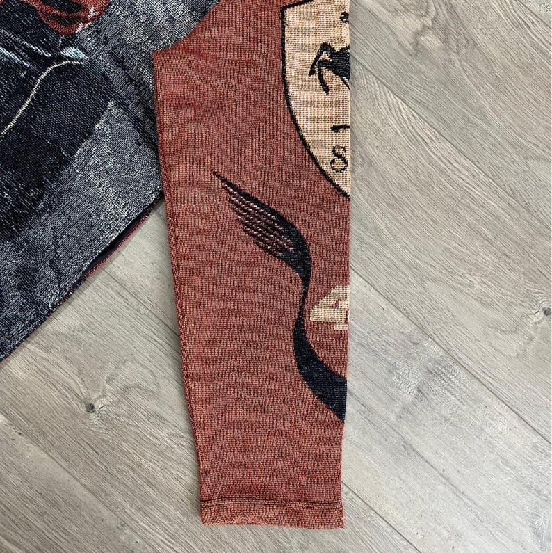 May include: A section of a woven textile featuring a shield with a black horse, the number 4, and other design elements. The textile has a reddish-brown background with black and beige details. The fabric appears to be a sleeve.