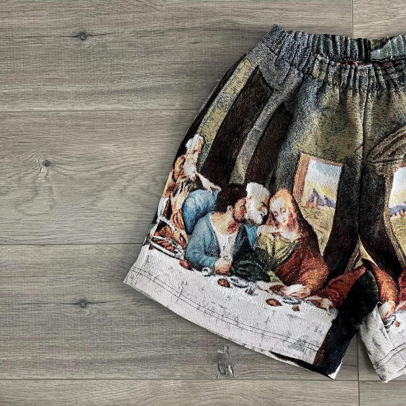 Op de afbeelding: Een zwart-witte short met een tapijt-achtige print van een schilderij dat een groep mensen laat zien die aan een tafel zitten. De print bevat verschillende kleuren, waaronder blauw, groen, rood en geel.