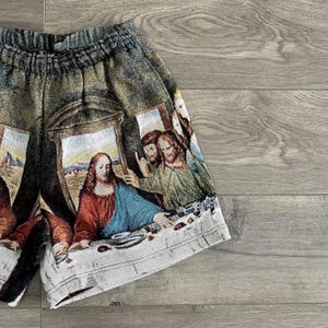Op de afbeelding: Een grijs en bruin short met een tapijt-achtig ontwerp met een sc&egrave;ne van mensen die rond een tafel zijn verzameld. De sc&egrave;ne doet denken aan Leonardo da Vinci's Het Laatste Avondmaal.