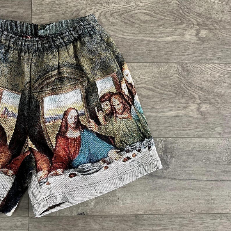 Op de afbeelding: Een grijs en bruin short met een tapijt-achtig ontwerp met een sc&egrave;ne van mensen die rond een tafel zijn verzameld. De sc&egrave;ne doet denken aan Leonardo da Vinci's Het Laatste Avondmaal.