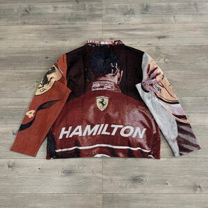 May include: A woven jacket featuring a portrait of a person with the word "HAMILTON" in white block letters. The jacket is primarily burgundy with accents of brown, tan, and cream. The Ferrari logo is visible on the jacket.