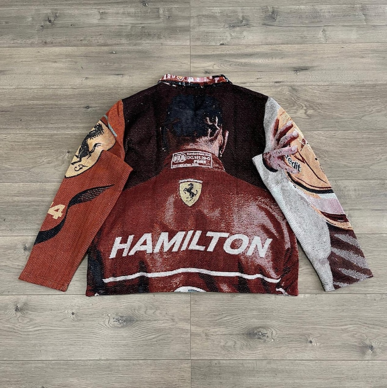 May include: A woven jacket featuring a portrait of a person with the word "HAMILTON" in white block letters. The jacket is primarily burgundy with accents of brown, tan, and cream. The Ferrari logo is visible on the jacket.