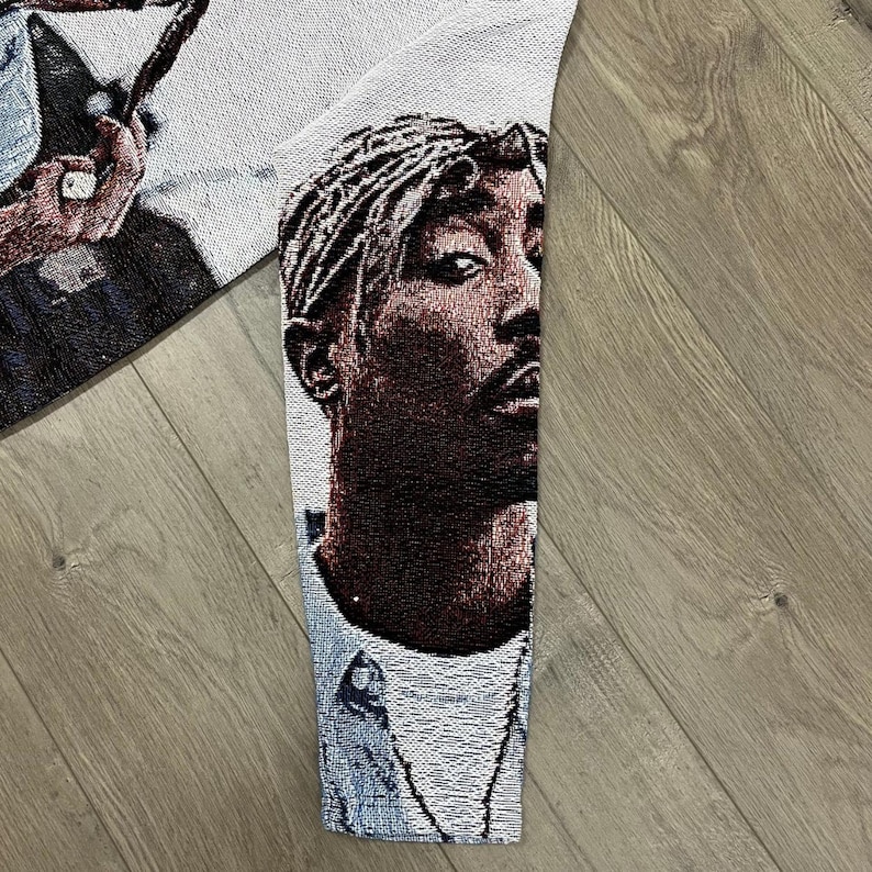 Op de afbeelding: Een geweven wandtapijt met een portret van Tupac Shakur. De afbeelding is in bruin, zwart en wit, met gedetailleerd weefwerk. Dit wandtapijt is waarschijnlijk een decoratief item, zoals een wandkleed of een sprei.