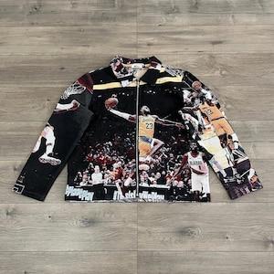 May include: Black zip-up jacket featuring a basketball game scene with players in action. The jacket has a collared neckline and long sleeves. The design includes the number 23 and the word "Blazers". The text "Play Play" and "#thesistheway" are also visible.