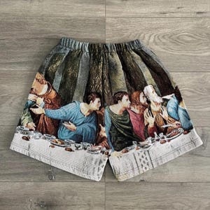 Op de afbeelding: Witte katoenen shorts met een kleurrijke print van een schilderij dat een groep mensen aan een tafel laat zien. De print bevat een verscheidenheid aan kleuren, waaronder rood, blauw, groen en geel.