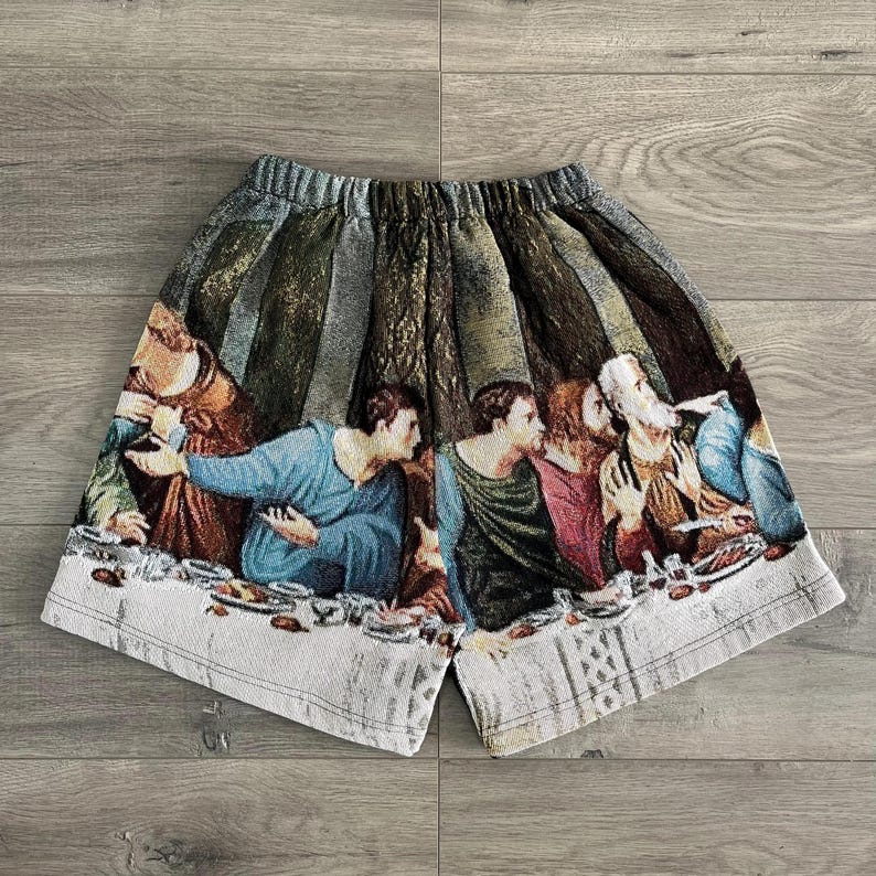 Op de afbeelding: Witte katoenen shorts met een kleurrijke print van een schilderij dat een groep mensen aan een tafel laat zien. De print bevat een verscheidenheid aan kleuren, waaronder rood, blauw, groen en geel.