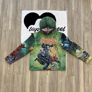 May include: Green hooded sweatshirt with a comic book print of Spider-Man and Venom fighting. The print features the text "The Amazing Spider-Man".