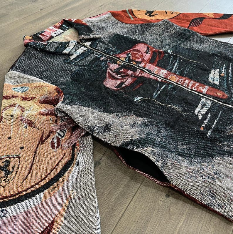 May include: A tapestry jacket with a full-length zipper and a collared neckline. The jacket features a complex design with shades of red, black, gray, and tan. The design includes a car and other graphic elements. The jacket has two pockets.