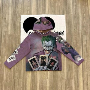 May include: Purple hoodie with a graphic print of the Joker from Batman. The Joker is holding four playing cards. The hoodie has a hood and long sleeves.
