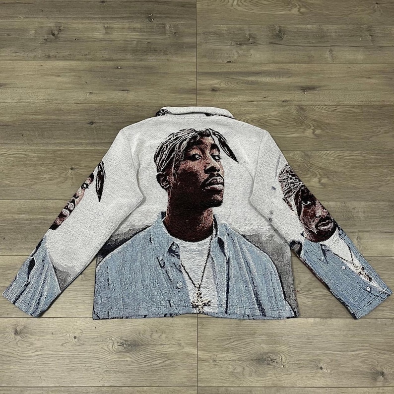 Op de afbeelding: Jas met lange mouwen met een portret van een persoon met een bandana en een kruisvormige hanger. De jas is voornamelijk wit met een lichtblauw shirtontwerp en portretdetails.