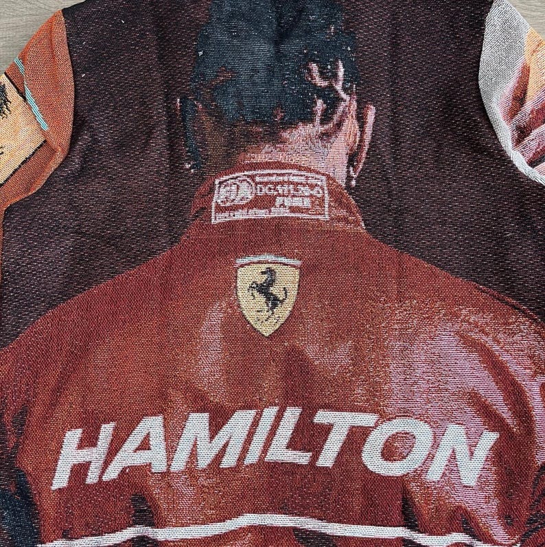 May include: A tapestry featuring a close-up of a red jacket with the name "HAMILTON" in white block letters. The jacket has a Ferrari logo and a rectangular patch with text. The background is a woven pattern.