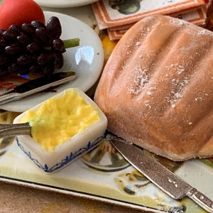 May include: A miniature table setting with a white plate, a small dish of yellow butter, a loaf of bread, a bunch of dark purple grapes, and two silver knives.
