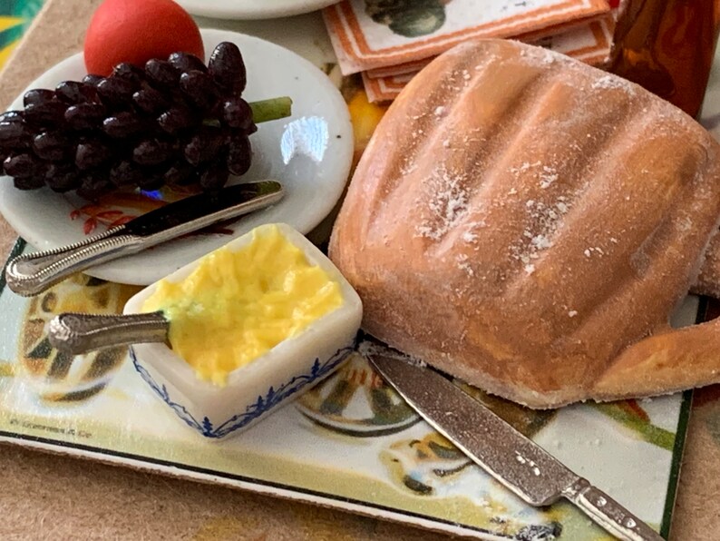 May include: A miniature table setting with a white plate, a small dish of yellow butter, a loaf of bread, a bunch of dark purple grapes, and two silver knives.