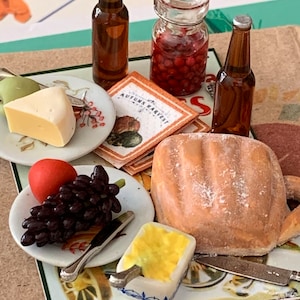 May include: A miniature table setting with a loaf of bread, a plate of grapes and a red ball, a plate of cheese and a green apple, a jar of red berries, two brown glass bottles, and a small dish of yellow butter. The table is decorated with a vintage-style placemat with the words "Autumn Harvest".