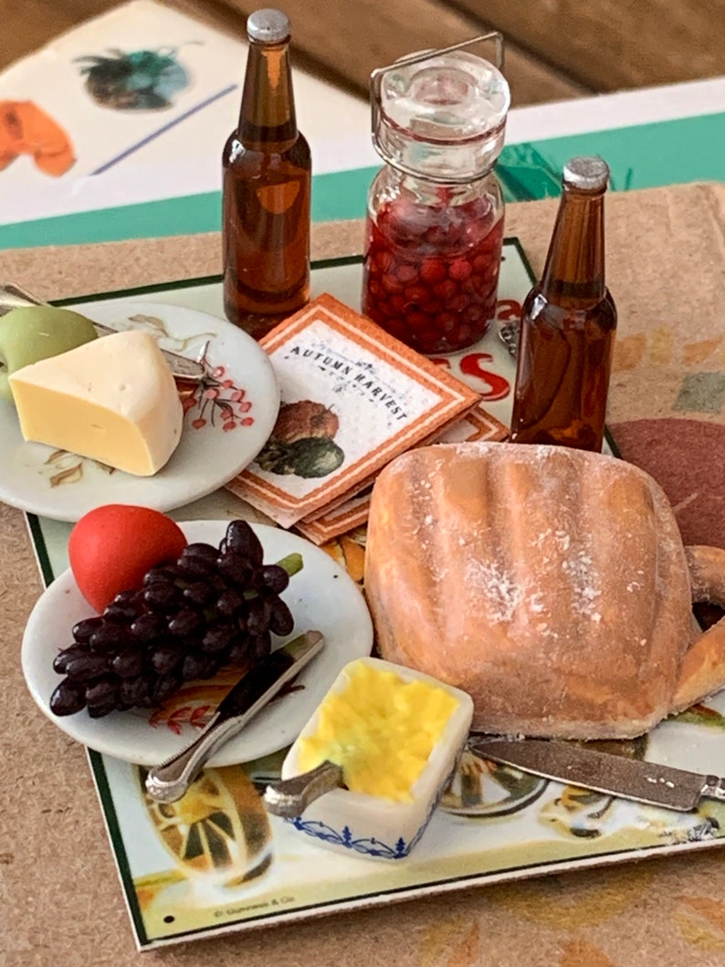 May include: A miniature table setting with a loaf of bread, a plate of grapes and a red ball, a plate of cheese and a green apple, a jar of red berries, two brown glass bottles, and a small dish of yellow butter. The table is decorated with a vintage-style placemat with the words "Autumn Harvest".