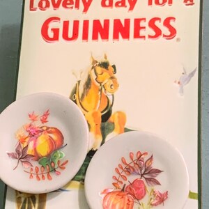 May include: Two white ceramic plates with a fall-themed design. The plates feature a pumpkin, leaves, and berries. The plates are displayed in front of a vintage Guinness sign that reads "Lovely day for a GUINNESS."