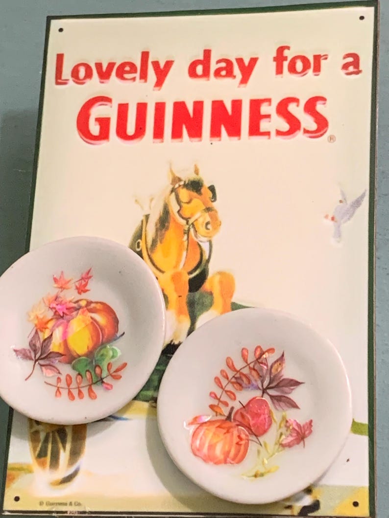 May include: Two white ceramic plates with a fall-themed design. The plates feature a pumpkin, leaves, and berries. The plates are displayed in front of a vintage Guinness sign that reads "Lovely day for a GUINNESS."