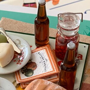 May include: A miniature picnic scene with a plate of cheese and an apple, two brown glass bottles, a jar of red cherries, and a napkin with the text "Autumn Harvest".