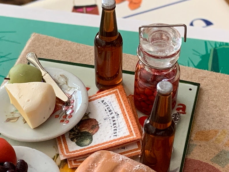May include: A miniature picnic scene with a plate of cheese and an apple, two brown glass bottles, a jar of red cherries, and a napkin with the text "Autumn Harvest".