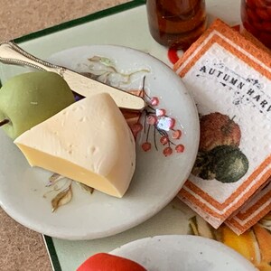 May include: A white ceramic plate with a green apple, a wedge of cheese, and a silver knife. The plate has a floral design with pink berries. The text "Autumn Harvest" is visible on a brown and white napkin.