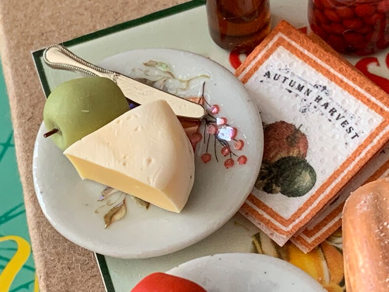 May include: A white ceramic plate with a green apple, a wedge of cheese, and a silver knife. The plate has a floral design with pink berries. The text "Autumn Harvest" is visible on a brown and white napkin.