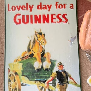 May include: A vintage metal sign with a green border and white background. The sign features a red and white illustration of a horse pulling a cart. The text on the sign reads "Lovely day for a GUINNESS".
