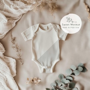 Pode incluir: Um macacão branco para bebê com um padrão xadrez cinza e branco na metade inferior. O macacão está sobre um fundo de tecido bege com flores secas e vegetação.