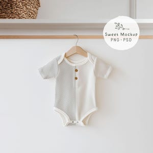 May include: A baby bodysuit with a two-tone design, half in a grey chequered pattern and half in solid white. The short-sleeved onesie has wooden buttons and is hanging on a wooden hanger. The overall theme is baby clothing.