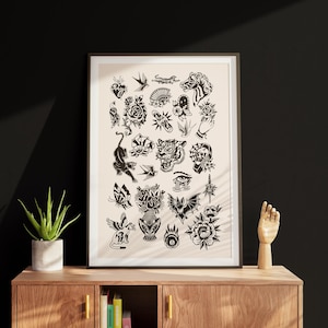 May include: A framed art print displaying a series of black and white tattoo-style illustrations. The artwork showcases traditional tattoo imagery, including a panther, flowers, and birds. The print is presented on a wooden cabinet, alongside a potted plant and a wooden hand sculpture.